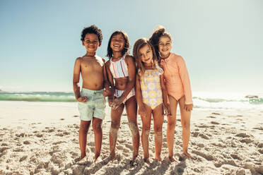 Enjoying the beach with friends. Four young children smiling cheerfully while standing together at the beach. Group of adorable little kids having fun together during summer vacation. - JLPSF09454