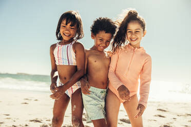 Unbekümmerte kleine Kinder lachen fröhlich, während sie am Strand mit Sand bedeckt sind. Gruppe von drei entzückenden kleinen Kindern, die in den Sommerferien zusammen Spaß haben. - JLPSF09458