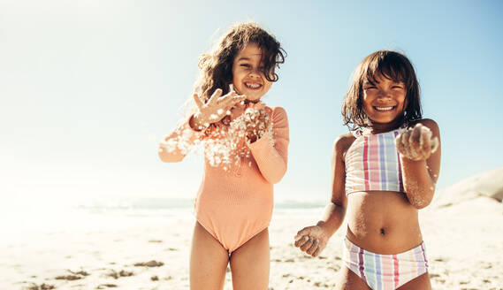 Fröhliche kleine Mädchen, die fröhlich lächelnd mit dem Sand am Strand spielen. Zwei entzückende kleine Kinder, die Spaß haben und sich in den Sommerferien amüsieren. - JLPSF09462