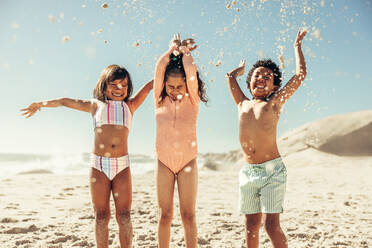Eine Gruppe von drei glücklichen Kindern, die ihren Sommerurlaub an einem sonnigen Strand genießen, hat Spaß daran, Sand in die Luft zu werfen. - JLPSF09463