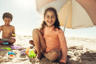 Happy young girl smiling at the camera while playing with her toys on beach sand. Cheerful little girl having a good time with her friends during summer vacation. - JLPSF09469