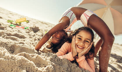 Cheerful little girls having fun together on beach sand. Two creative young friends smiling happily while playing with each other at the beach. Adorable kids enjoying their summer vacation. - JLPSF09471
