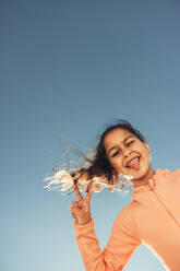 Niedliches kleines Mädchen, das das Friedenszeichen zeigt, während es die Zunge herausstreckt. Bezauberndes junges Mädchen, das im Sommer am Strand Spaß hat. Glückliches kleines Mädchen, das mit dem Himmel im Hintergrund steht. - JLPSF09473