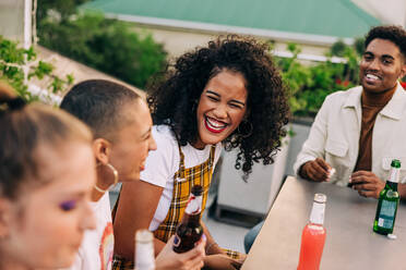 Happy young woman laughing with her friends outdoors. Group of happy young people having some cold beers while hanging out on a rooftop. Cheerful friends having a good time on the weekend. - JLPSF09482