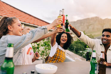 Cheers to the weekend. Group of happy young friends making a toast with cold beers while hanging out together on a rooftop. Cheerful young friends laughing and having a good time outdoors. - JLPSF09483
