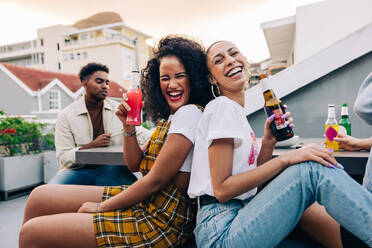 Best friends enjoying cold drinks on the weekend. Two happy young women having a good time while hanging out with their friends on a rooftop. Two female friends laughing and holding beer bottles. - JLPSF09484