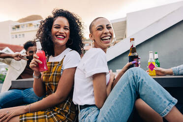 Freunde, die am Wochenende ein paar kalte Getränke genießen. Zwei glückliche junge Frauen, die sich mit ihren Freunden auf einem Dach amüsieren. Zwei fröhliche Freundinnen, die lächeln und Bierflaschen halten. - JLPSF09485