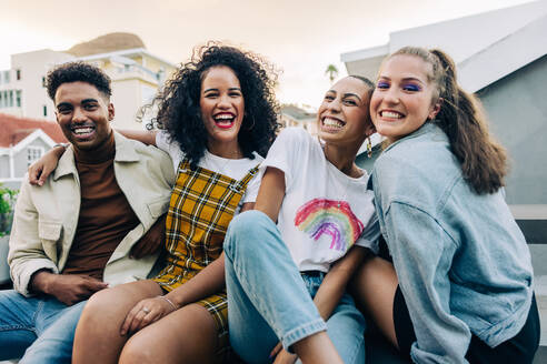 Friends smiling at the camera while sitting on a rooftop. Group of cheerful young friends enjoying hanging out together on the weekend. Four happy friends having a good time together. - JLPSF09487