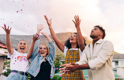 Multicultural friends enjoying a party on a rooftop. Group of vibrant friends cheering and throwing confetti into the air. Four happy friends celebrating and having fun on the weekend. - JLPSF09490