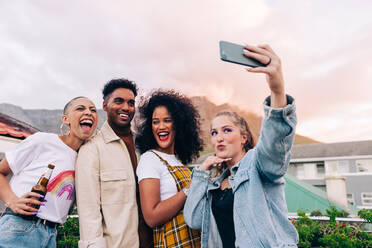Friends taking a group selfie outdoors. Four happy friends posing for a selfie while partying together on the rooftop. Group of multicultural friends having a good time together on the weekend. - JLPSF09497