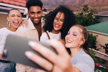 Freunde lächeln für ein Gruppen-Selfie auf einem Dach. Vier glückliche Freunde, die ein Foto mit einem Fotohandy machen. Eine Gruppe multikultureller Freunde, die am Wochenende eine gute Zeit miteinander verbringen. - JLPSF09499