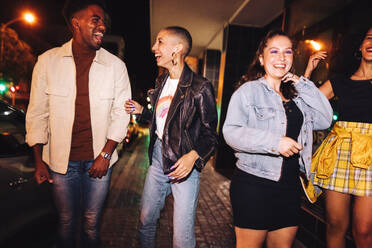 Eine Gruppe multikultureller Freunde, die zusammen in der Stadt ausgehen. Fröhliche Freunde, die lachen, während sie nachts im Freien spazieren gehen. Lebhafte junge Leute, die sich am Wochenende gut amüsieren. - JLPSF09525