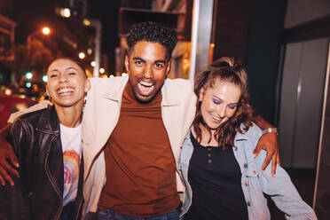 Drei junge Freunde, die sich nachts in der Stadt amüsieren. Eine Gruppe lebhafter junger Leute, die fröhlich lachen, während sie zusammen spazieren gehen. Multikulturelle Freunde, die am Wochenende zusammen ausgehen. - JLPSF09527