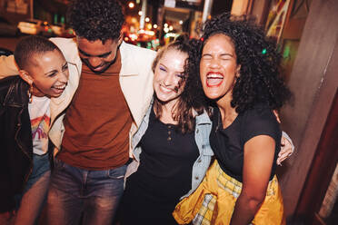 Friday night fun. Group of vibrant young people laughing joyfully while walking together in the city at night. Four multicultural friends going out together on the weekend. - JLPSF09529