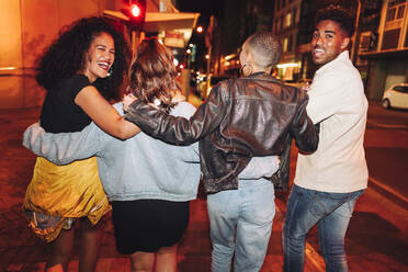 Rückansicht einer Gruppe von Freunden, die nachts durch die Stadt spazieren. Eine Gruppe junger Leute lacht fröhlich und umarmt sich. Multikulturelle Freunde, die am Wochenende zusammen ausgehen. - JLPSF09530