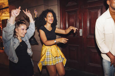 Fröhlich tanzende Freundinnen in der nächtlichen Stadt. Eine Gruppe lebhafter junger Frauen, die lachen und sich amüsieren, während sie gemeinsam im Freien spazieren gehen. Multikulturelle Freunde, die am Wochenende ausgehen. - JLPSF09536
