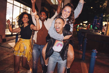 Cheerful young woman piggybacking her friend in the city. Girlfriends cheering and having fun while going out with their friends at night. Group of vibrant friends hanging out together on the weekend. - JLPSF09554