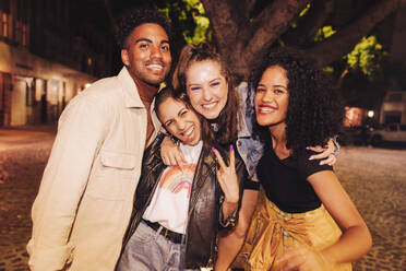 Group of multicultural friends smiling at the camera cheerfully. Happy young friends having a good time while hanging out together outdoors. Vibrant young people enjoying a weekend night in the city. - JLPSF09576