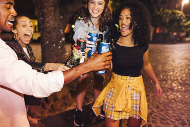 Frohes neues Jahr. Lebhafte Freunde stoßen mit verschütteten Bierdosen an, während sie nachts im Freien stehen. Eine Gruppe ekstatischer junger Leute, die Silvester mit kalten Getränken feiern. - JLPSF09579