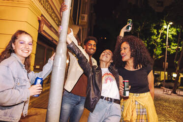 Enjoying cold drinks in the city. Group of cheerful young friends smiling happily while holding beer cans. Multicultural young people having a good time together on the weekend. - JLPSF09581