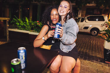 Freundinnen, die in einem Restaurant im Freien Bierdosen halten. Zwei fröhliche junge Frauen, die bei einem Mädelsabend glücklich lachen. Freunde, die sich am Wochenende amüsieren. - JLPSF09589