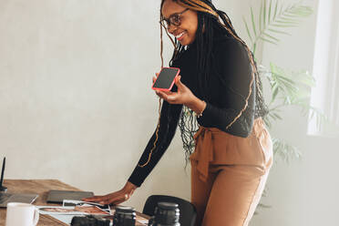 Kreative Freiberuflerin, die einen Anruf in ihrem Büro zu Hause entgegennimmt. Glückliche junge Frau, die lächelt, während sie an einem Schreibtisch mit Fotoausrüstung steht. Fotografin, die mit ihren Kunden kommuniziert. - JLPSF09616