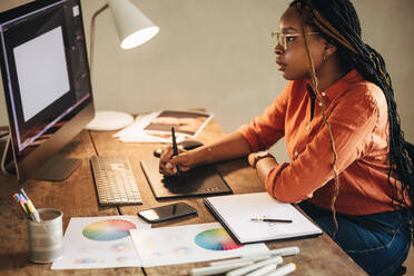 Freiberuflerin, die an ihrem Schreibtisch an einem neuen Design arbeitet. Konzentrierte junge Grafikdesignerin, die mit einem Stylus-Stift auf einem digitalen Tablet zeichnet. Kreative junge Frau, die in ihrem Heimbüro arbeitet. - JLPSF09633
