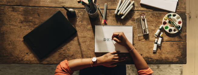 Woman drawing the word ''DESIGN'' while sitting at her creative workstation. High angle view of a female graphic designer working on a new project. Anonymous young freelancer working in her home office. - JLPSF09638