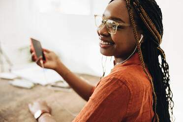 Unbeschwerte Freiberuflerin, die Musik auf ihrem Smartphone hört. Glückliche junge Frau, die lächelnd in ihrem Heimbüro sitzt. Junge Grafikdesignerin, die an einem kreativen Arbeitsplatz arbeitet. - JLPSF09644