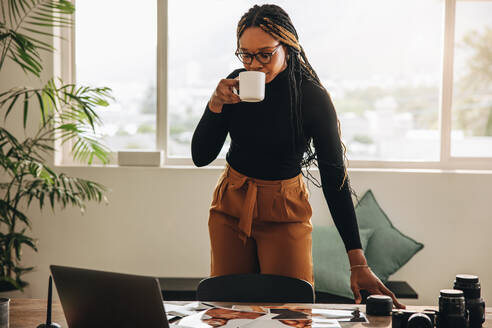 Selbstständige junge Frau, die in ihrem Büro zu Hause Kaffee trinkt. Kreative junge Frau, die an einem Schreibtisch mit Fotoausrüstung steht. Fotografin, die eine Kaffeepause macht, während sie an einem neuen Projekt arbeitet. - JLPSF09657