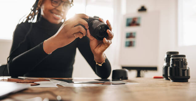 Frau hält eine DSLR-Kamera in ihrem Büro zu Hause. Kreative junge Fotografin lächelt fröhlich, während sie an ihrem Schreibtisch arbeitet. Freiberuflerin arbeitet an einem neuen fotografischen Projekt. - JLPSF09666