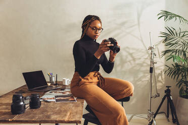 Kreative junge Fotografin, die eine DSLR-Kamera in ihrem Büro zu Hause hält. Künstlerische junge Frau, die ihre Kamera einrichtet, während sie an einem neuen Projekt arbeitet. Freiberuflerin, die oben auf ihrem Schreibtisch sitzt. - JLPSF09677