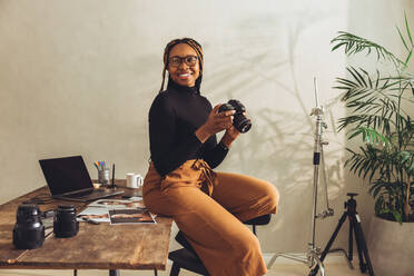 Glückliche Fotografin, die fröhlich lächelt, während sie eine DSLR-Kamera hält. Kreative junge Frau, die nachdenklich wegschaut, während sie an ihrem Schreibtisch sitzt. Freiberuflerin, die an einem neuen Projekt in ihrem Büro zu Hause arbeitet. - JLPSF09678