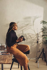 Künstlerin hält eine DSLR-Kamera in ihrem Büro zu Hause. Kreative junge Fotografin schaut nachdenklich, während sie auf ihrem Schreibtisch sitzt. Freiberuflerin arbeitet an einem neuen Projekt in ihrem Arbeitsbereich. - JLPSF09679