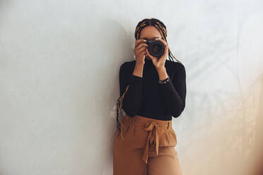 Capturing through a dslr camera. Young female photographer taking a picture while standing against a wall in her home office. Creative female freelancer working from home. - JLPSF09691