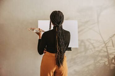 Kreative junge Frau hält eine Palette in der Hand, während sie vor einer Leinwand steht. Rückansicht einer Malerin, die in einem Kunstatelier über neue Ideen nachdenkt. Künstlerische Freiberuflerin bei der Arbeit an einem neuen Projekt. - JLPSF09695