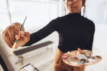 Junge Malerin, die mit einem Pinsel auf eine Leinwand zeichnet. Kreative junge Frau, die eine Palette hält, während sie an einem Gemälde arbeitet. Künstlerische Freiberuflerin, die in ihrem Heimatelier arbeitet. - JLPSF09698