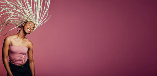 Afroamerikanische Frau mit langen geflochtenen Haaren, die mit geschlossenen Augen tanzt und ihre geflochtenen Haare in der Luft fliegen lässt, auf rosa Hintergrund. - JLPSF09718