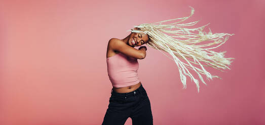 Porträt einer glücklichen Frau, die auf einem rosafarbenen Hintergrund tanzt und dabei ihre langen geflochtenen Haare fliegen lässt. - JLPSF09720