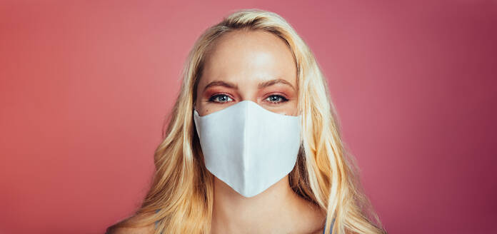 Close-up of a woman with blonde hair wearing face mask on pink background. Female with eye makeup wearing protective face mask looking at camera. - JLPSF09725