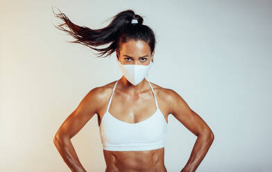 Porträt einer selbstbewussten Sportlerin mit Schutzmaske. Muskulöse Frau in Sportkleidung und mit Gesichtsmaske, die Hände in die Hüften gestemmt, vor weißem Hintergrund. - JLPSF09739