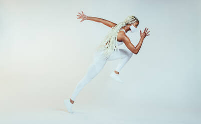 Sportliche Frau mit Schutzmaske, die über einen weißen Hintergrund läuft. Ganzkörperaufnahme einer gesunden afrikanischen Frau beim Sprinten. - JLPSF09759