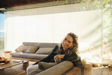 Glückliche reife Frau lächelt in die Kamera, während sie mit einer Tasse Tee auf einer Couch sitzt. Fröhliche ältere Frau entspannt sich in ihrem Wohnzimmer zu Hause. - JLPSF09795