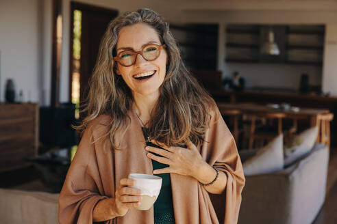 Beautiful senior woman smiling at the camera while standing with a cup of tea in her hand. Cheerful senior woman enjoying a happy retirement at home. - JLPSF09805