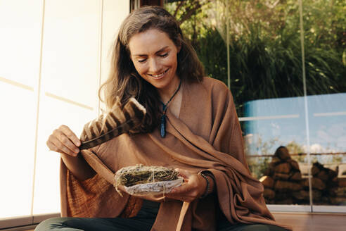 Happy senior woman smiling while holding a feather and a sage smudge stick. Mature woman performing a purifying and cleansing ceremony at home. Cheerful woman practicing alternative medicine. - JLPSF09816