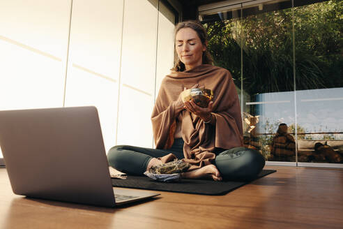 Ayurvedic healer meditating with a singing bowl and sage during an online holistic class. Woman performing a self-healing and purifying ceremony at home. Senior woman taking care of her ageing body. - JLPSF09825