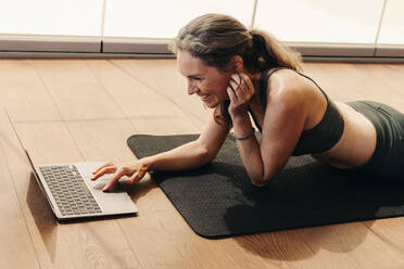 Ältere Frau lächelt, während sie an einem Online-Fitnesskurs auf einem Laptop teilnimmt. Glückliche Frau, die einem Online-Yoga-Tutorial zu Hause folgt. Fröhliche ältere Frau, die auf einer Übungsmatte liegt. - JLPSF09829