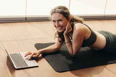 Ältere Frau, die in die Kamera lächelt, während sie an einer virtuellen Yogastunde auf einem Laptop teilnimmt. Glückliche Frau, die einem Online-Workout-Tutorial zu Hause folgt. Fröhliche ältere Frau, die auf einer Übungsmatte liegt. - JLPSF09830