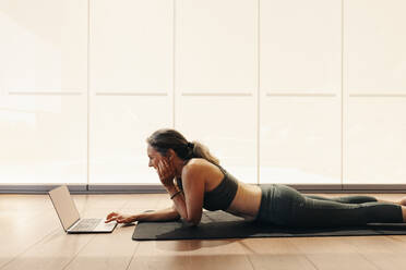 Happy senior woman smiling while joining an online yoga class on a laptop. Woman following a virtual fitness tutorial at home. Cheerful mature woman lying on an exercise mat. - JLPSF09831