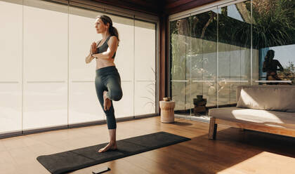 Ältere Frau, die während einer Yogasitzung die Baumstellung übt. Ältere Frau, die auf einer Übungsmatte eine Gleichgewichts-Asana macht. Aktive Frau, die zu Hause ein gesundes Trainingsprogramm absolviert. - JLPSF09836